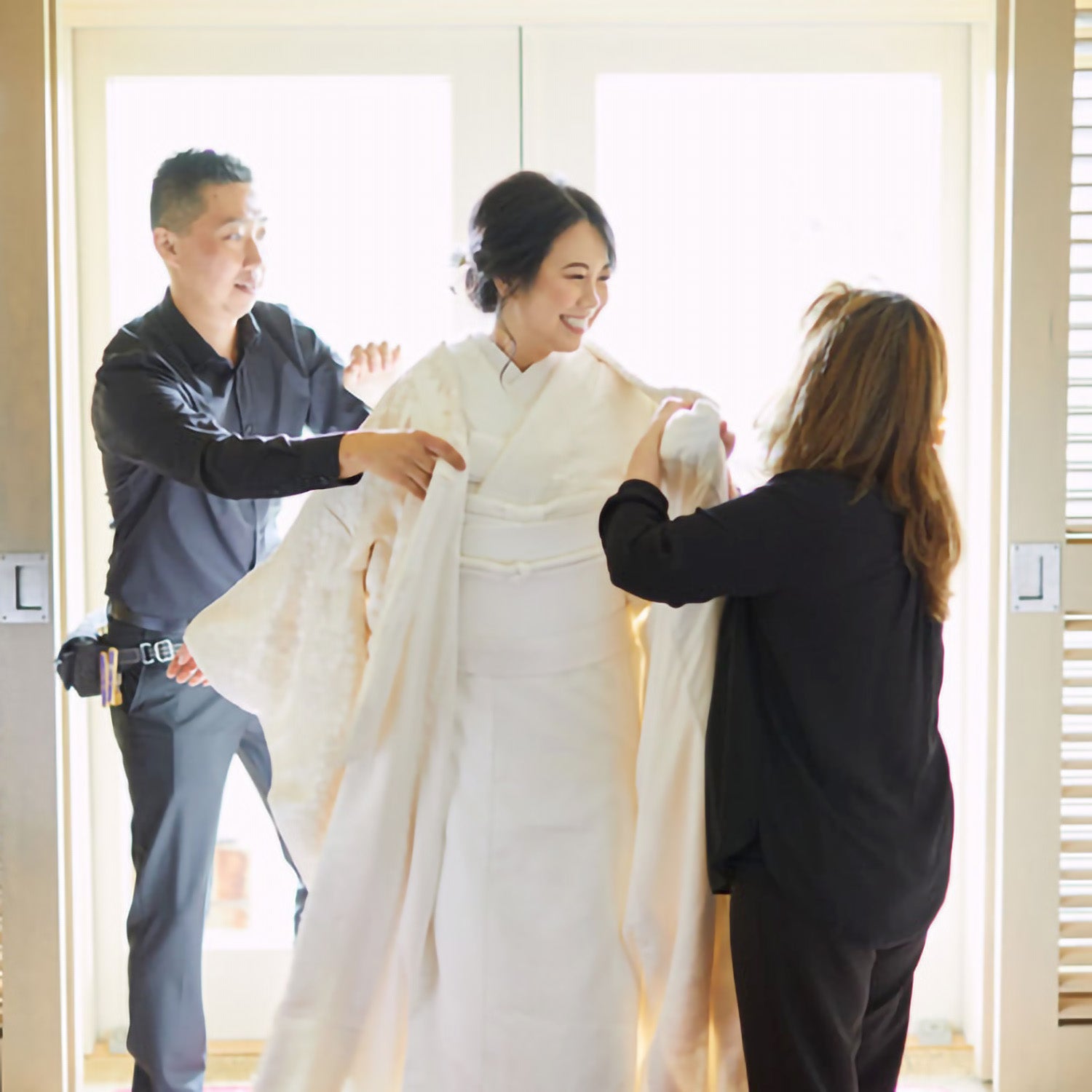 KIMONO SK founders Sueko Oshimoto and Kentaro Terauchi assist a smiling bride with her traditional white Japanese wedding kimono in a beautifully sunlit room.