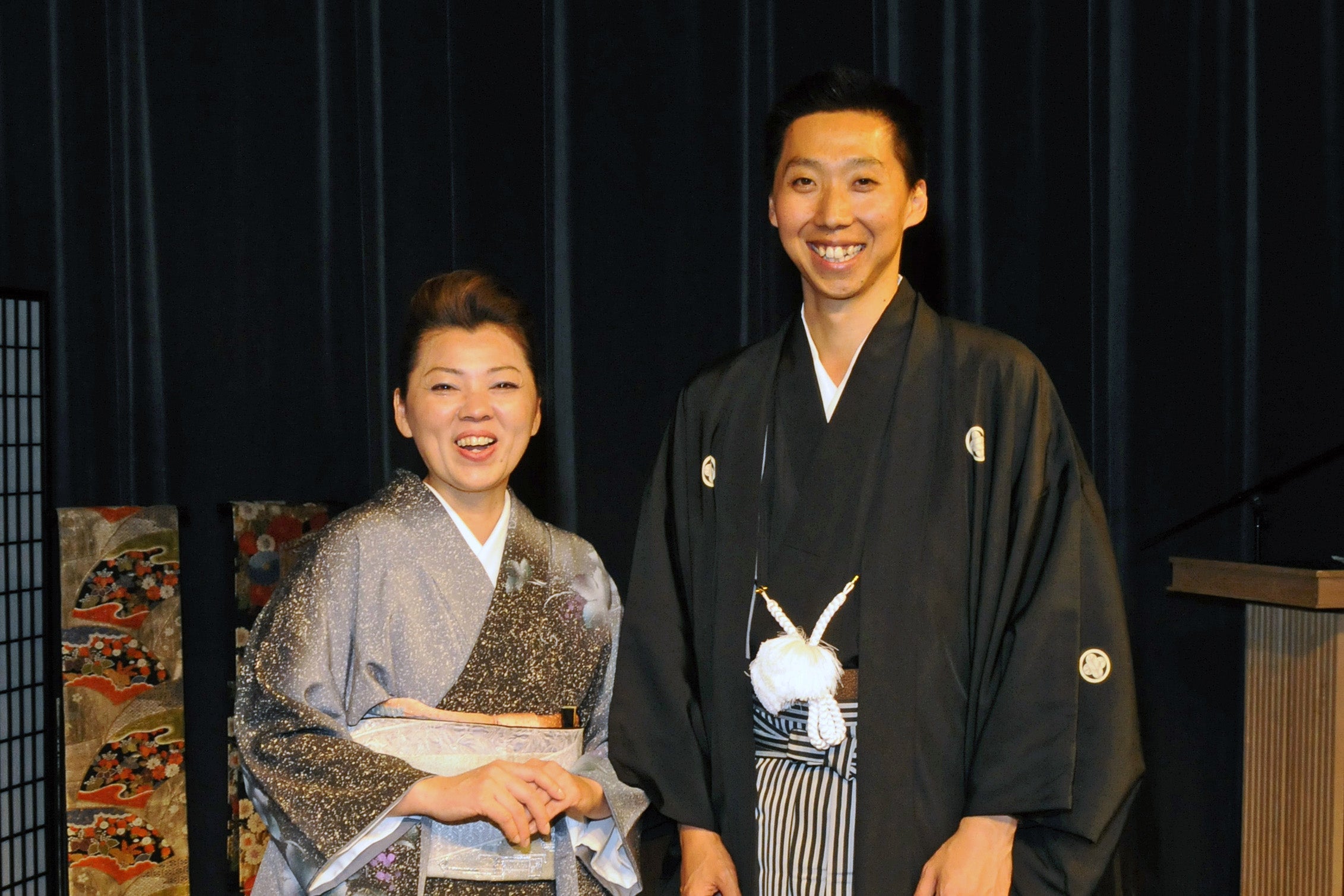 Two people in traditional Japanese attire standing against a dark curtain.
