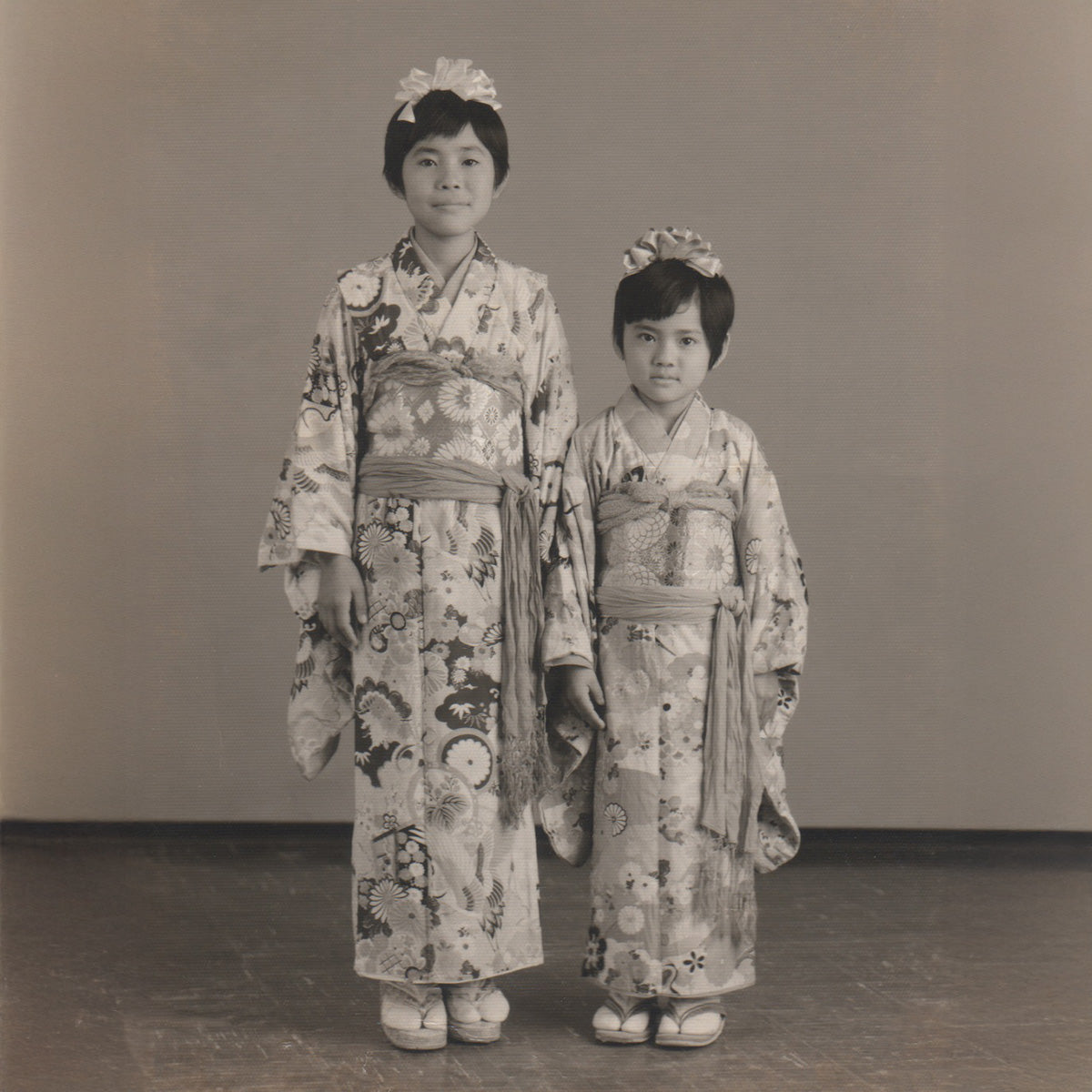 A nostalgic black and white photo from 1974 showing a young Sueko Oshimoto, founder of KIMONO SK, standing with her sister, both dressed in traditional Japanese kimonos, marking the origin of her lifelong passion.