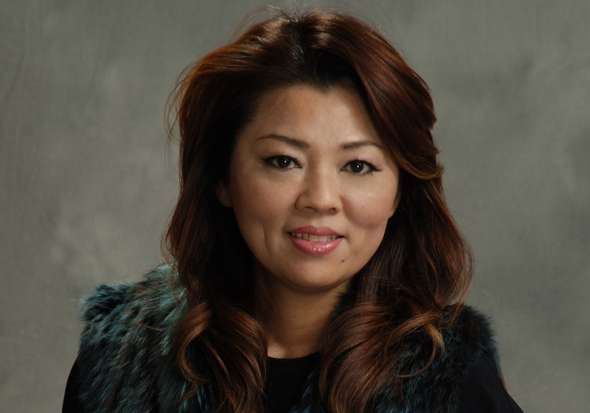 Sueko Oshimoto wearing a dark coat with a fur vest against a gray background